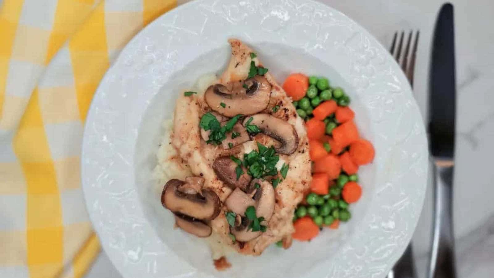A white plate with grilled chicken topped with mushrooms and herbs, served over mashed potatoes. It is accompanied by a side of peas and carrots. A yellow checkered cloth and utensils are next to the plate.