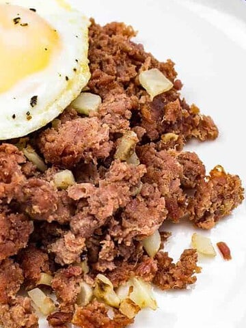 A plate with a sunny-side-up egg and a serving of corned beef hash. The egg has a runny yolk, and the hash contains crumbled corned beef mixed with small pieces of onion. The dish is on a white plate.