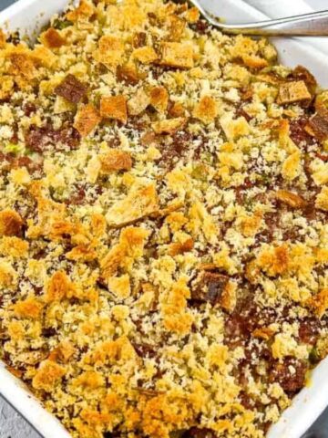 A square white baking dish holds a freshly baked dessert with a golden-brown crumbly topping. A serving spoon rests on the edge of the dish. The dish is on a white surface with floral-patterned and plain napkins nearby.