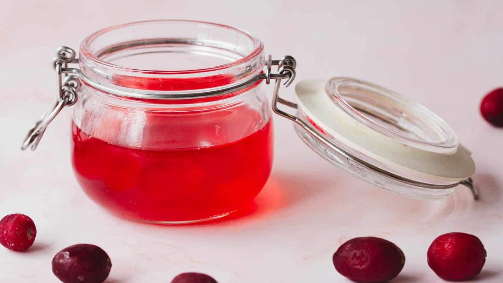 A glass jar filled with red jelly is shown with the lid resting open beside it. The jar is placed on a light surface, and a few cranberries are scattered around.