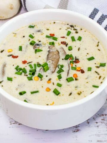 A white ceramic bowl filled with creamy mushroom soup, a staple among Grandma meals, is garnished with chopped chives and red pepper flakes. The bowl rests on a rustic white wooden surface beside a striped cloth napkin and a few whole mushrooms, evoking nostalgic flavors.