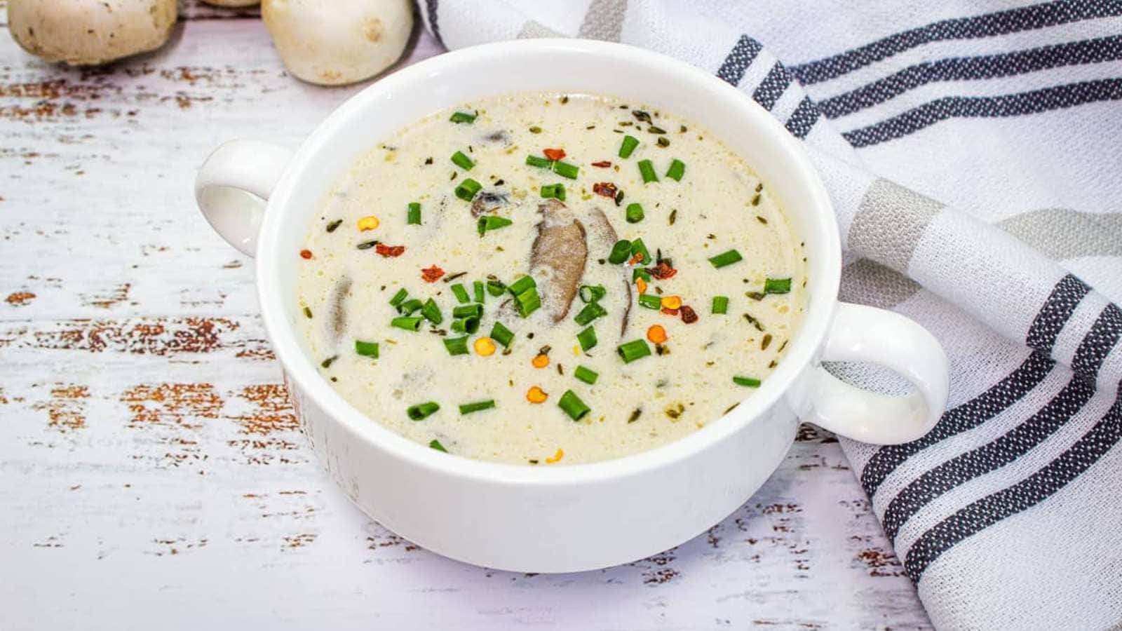 A white ceramic bowl filled with creamy mushroom soup, a staple among Grandma meals, is garnished with chopped chives and red pepper flakes. The bowl rests on a rustic white wooden surface beside a striped cloth napkin and a few whole mushrooms, evoking nostalgic flavors.