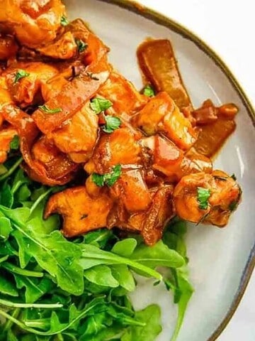 A plate with chicken pieces in a reddish sauce and a side of arugula. A fork is visible in the background.