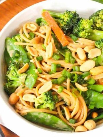 A bowl of noodles with broccoli, carrot strips, snow peas, and peanuts, garnished with green onions. The bowl is on a white napkin on a wooden plate, with a fork and green onions alongside.