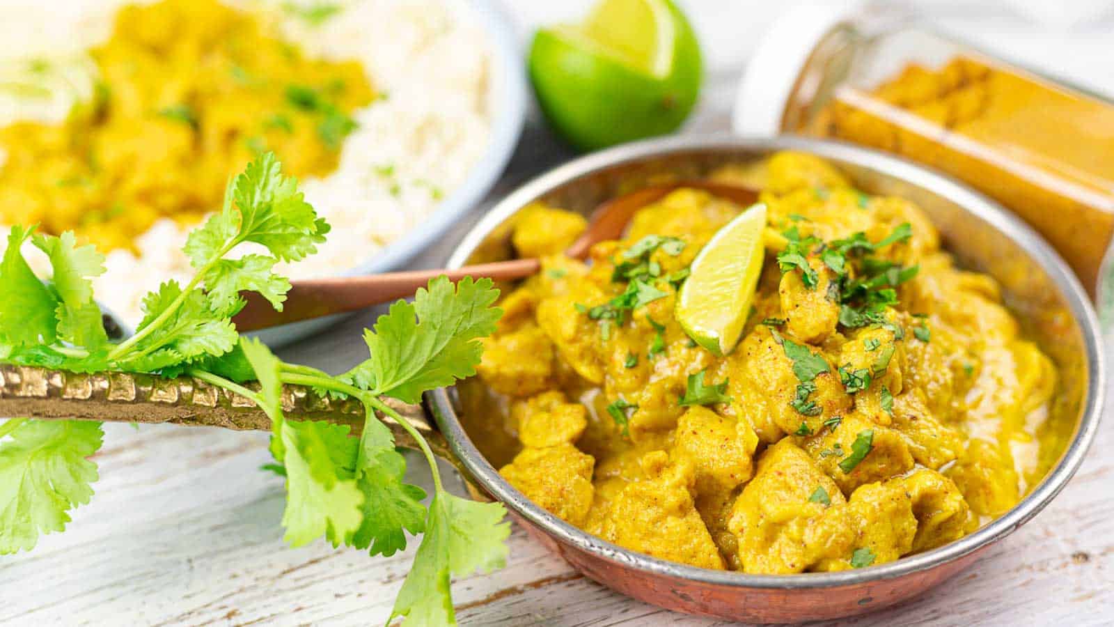 A bowl of curry featuring chunks of meat in a creamy yellow sauce, garnished with a lime wedge and cilantro. Nearby, a bowl of rice and a lime, with a sprinkle of fresh cilantro and spices in a jar.