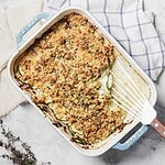 A rectangular baking dish holds a cheesy zucchini casserole with a crumbly topping. Resting on a white surface next to a folded blue-checkered cloth and sprigs of fresh thyme, this delightful dish invites you to dig in with the serving spatula placed on top.
