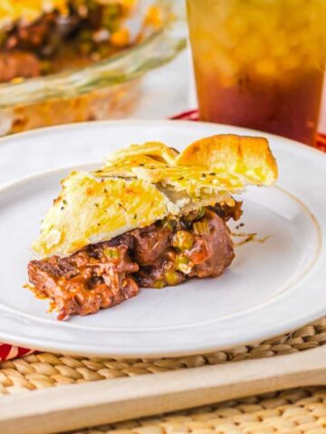 Slice of savory pie with a golden crust, served on a white plate. The filling includes meat and vegetables. The plate rests on a red patterned cloth, with a wooden spoon nearby. A glass of iced beverage and the remaining pie are in the background.
