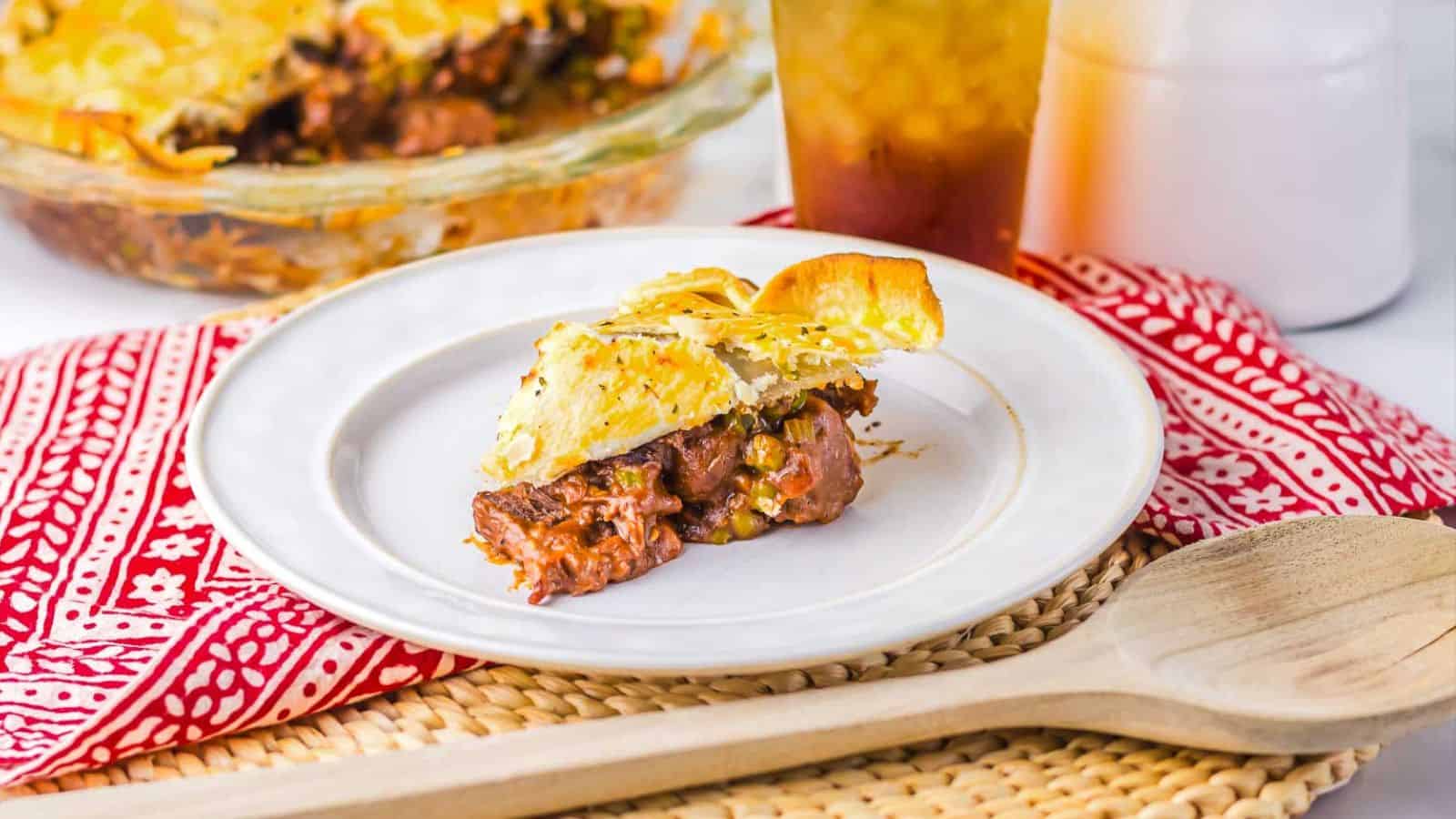 A close-up of a beef and vegetable pie with a golden, flaky crust. A triangular segment is removed, revealing chunks of beef and vegetables in a savory sauce inside the pie. The crust is lightly seasoned with herbs.