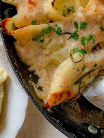 A skillet brimming with cheesy baked stuffed pasta shells draped in a creamy sauce, garnished with chopped parsley. A spoon rests invitingly in the skillet, while to the side a white plate awaits with more delectable dishes of stuffed shells on a light-colored surface.