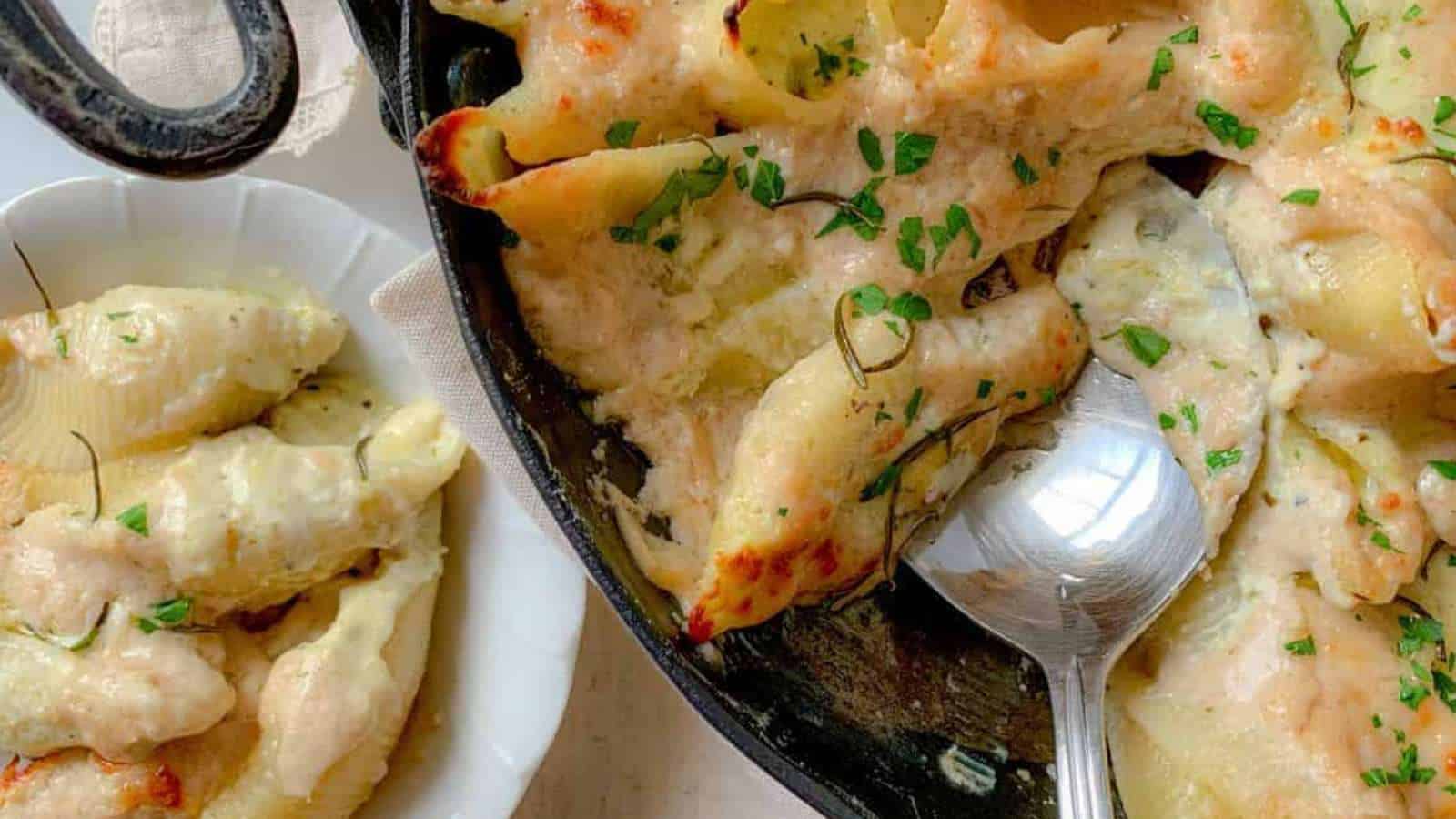A skillet brimming with cheesy baked stuffed pasta shells draped in a creamy sauce, garnished with chopped parsley. A spoon rests invitingly in the skillet, while to the side a white plate awaits with more delectable dishes of stuffed shells on a light-colored surface.