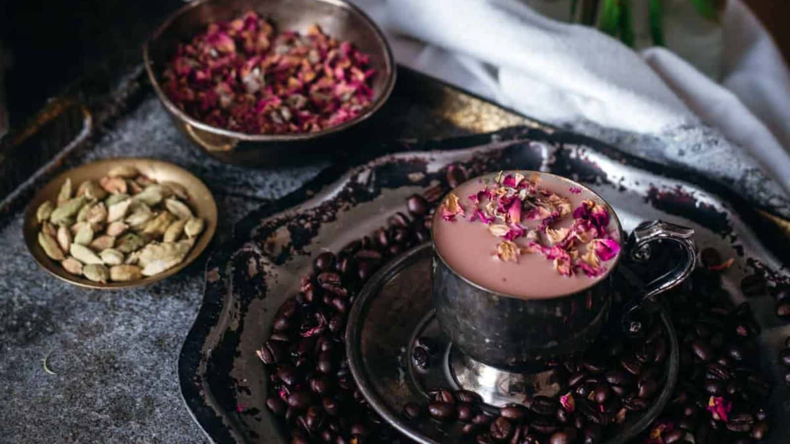 A cup filled with pinkish liquid topped with dried rose petals sits on a dark tray surrounded by coffee beans. Nearby, a small dish contains cardamom pods, and another bowl holds more dried rose petals. A white cloth is partially visible.
