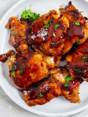 A white plate with glazed grilled chicken thighs garnished with fresh herbs. The chicken appears moist and coated with a dark, rich sauce. The plate is set on a light-colored surface, accompanied by a small green sprig for decoration.