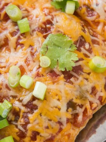 A close-up of a baked dish featuring enchiladas topped with melted cheese, chopped green onions, and cilantro. Some of the enchiladas have been cut to reveal a filling inside. The dish is in a glass baking tray.