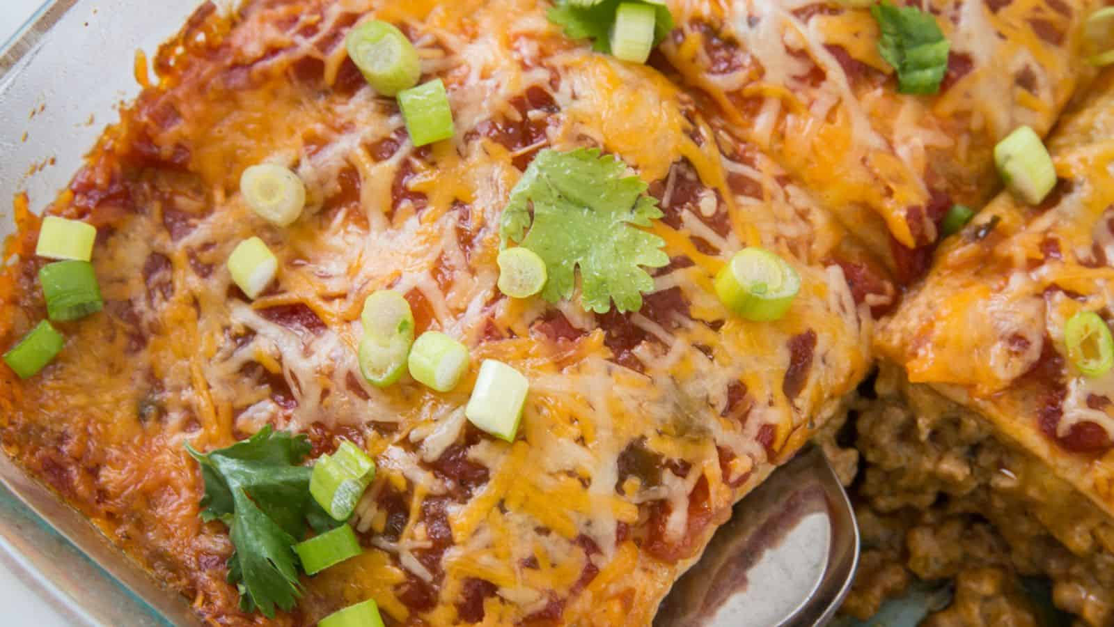 A close-up of a baked dish featuring enchiladas topped with melted cheese, chopped green onions, and cilantro. Some of the enchiladas have been cut to reveal a filling inside. The dish is in a glass baking tray.