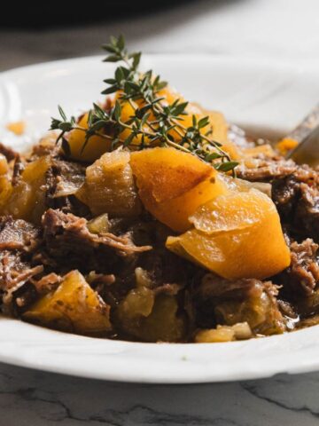 A white plate containing a stew made of tender shredded meat and chunks of potatoes, garnished with sprigs of fresh thyme. A spoon rests on the side of the dish, placed on a marble surface.