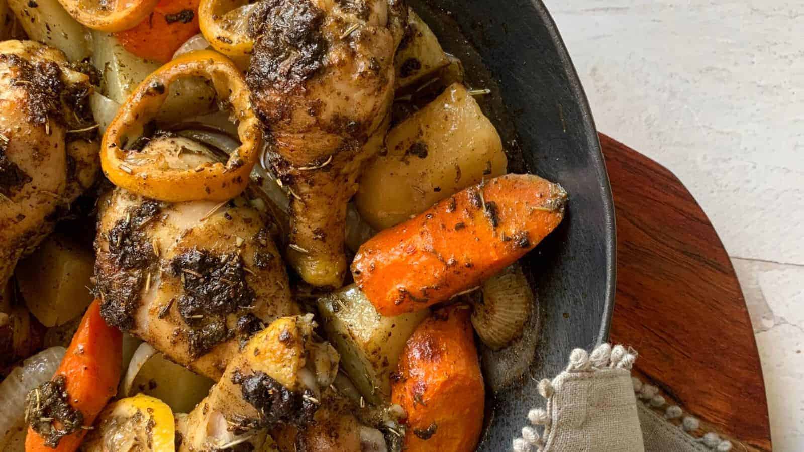 A black skillet filled with roasted chicken drumsticks, sliced carrots, potatoes, and lemon slices, seasoned with herbs. Perfect for a relaxing weekend meal, this no-stress recipe rests on a wooden surface with part of a cloth napkin visible.