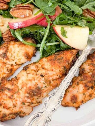 A white plate with three pieces of seasoned grilled chicken breast next to a salad. The salad contains arugula, sliced apples, and pecans. An ornate fork rests on the plate.