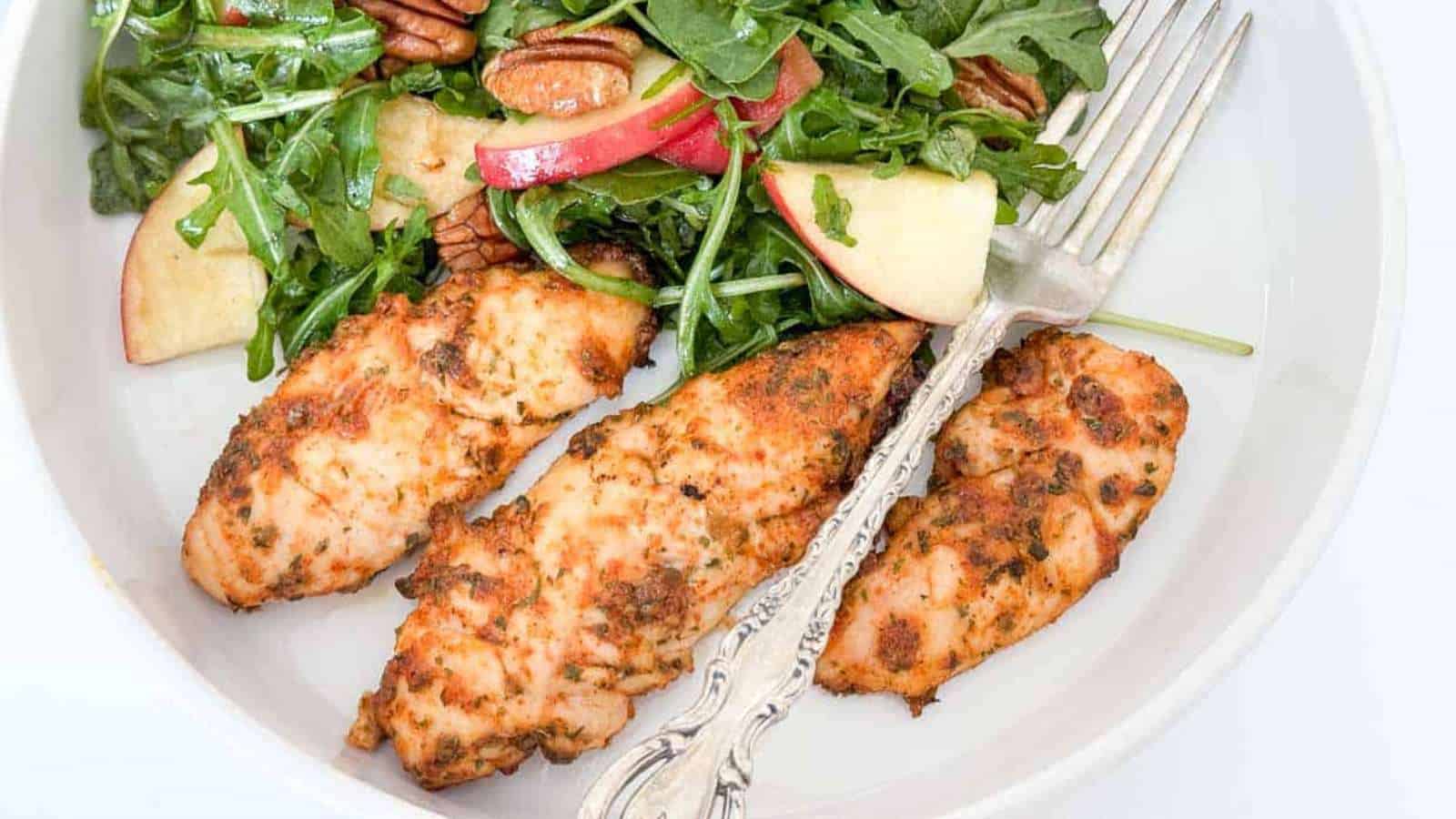 A white plate with three pieces of seasoned grilled chicken breast next to a salad. The salad contains arugula, sliced apples, and pecans. An ornate fork rests on the plate.