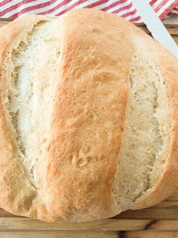 A round loaf of bread with a crusty exterior sits on a wooden cutting board. A serrated knife with a black handle is placed beside it. A red and white striped cloth is partially visible in the background.