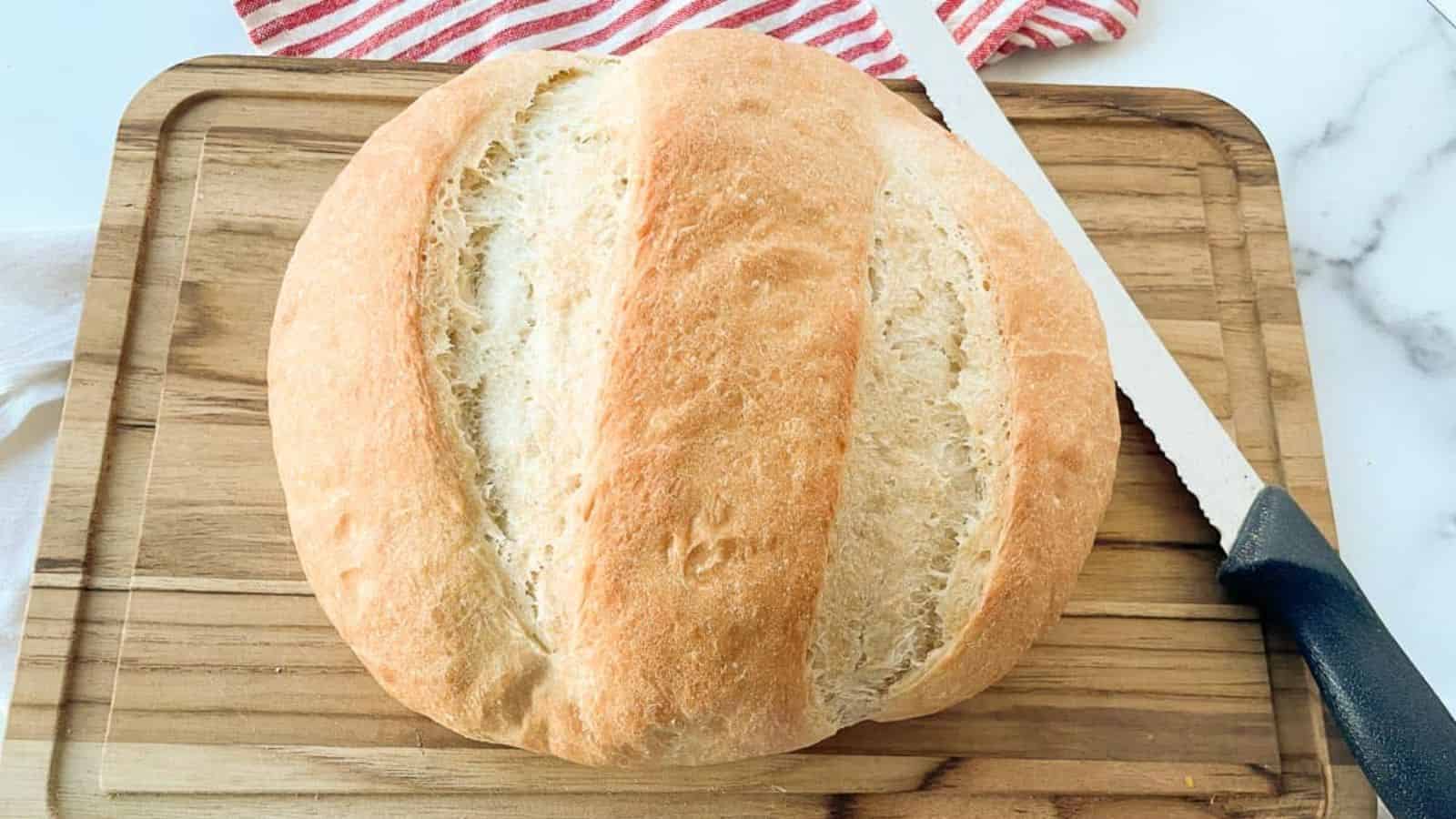 A loaf of peasant bread on a cutting baord with a knife with it.