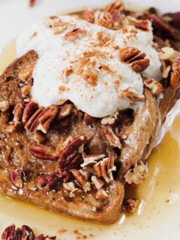 A slice of French toast on a white plate is topped with whipped cream, pecans, a dusting of cinnamon, and drizzled with syrup.