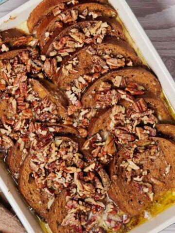 A rectangular baking dish filled with sliced brown bread, covered in a sauce and topped with chopped pecans. The dish is on a light wood surface next to a brown cloth and a small bowl containing a white creamy substance.