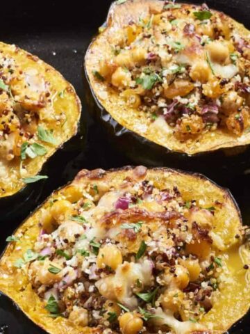 A cast iron skillet holds three halves of roasted acorn squash filled with a stress-free mixture of chickpeas, quinoa, herbs, and melted cheese. Perfect for impressing in-laws, a serving spoon rests on the skillet's edge.