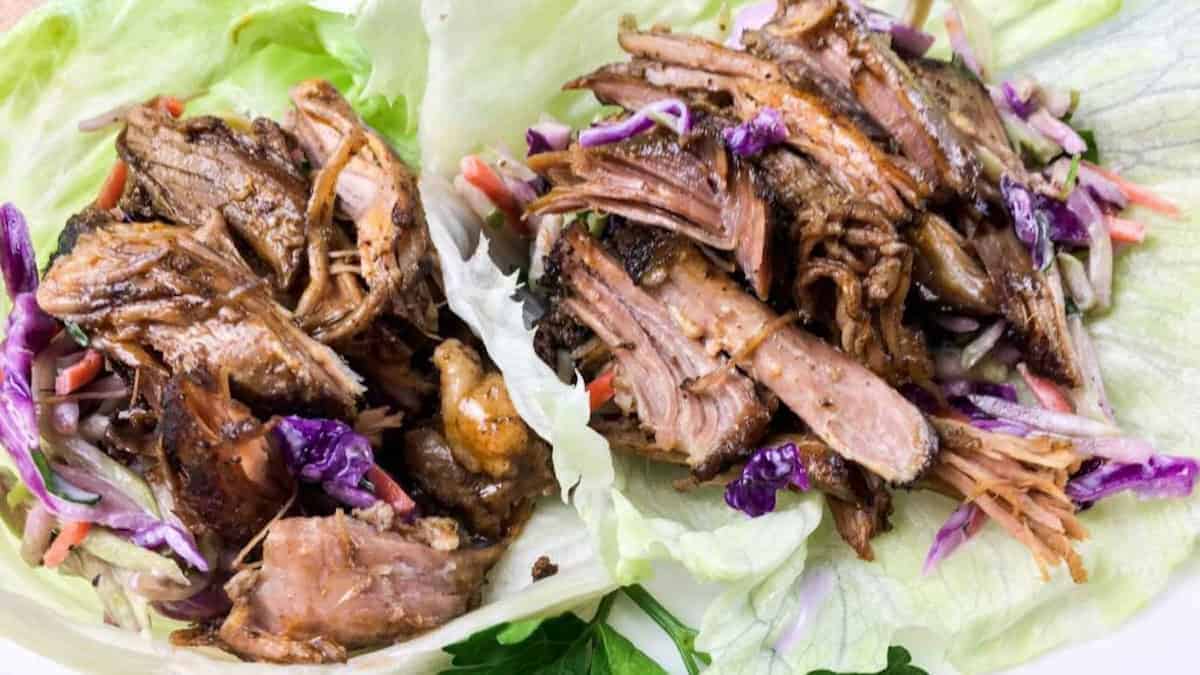 Two lettuce wraps filled with shredded meat and colorful slaw featuring purple cabbage and carrots. The meat appears tender and well-cooked, sitting atop crisp green lettuce leaves.