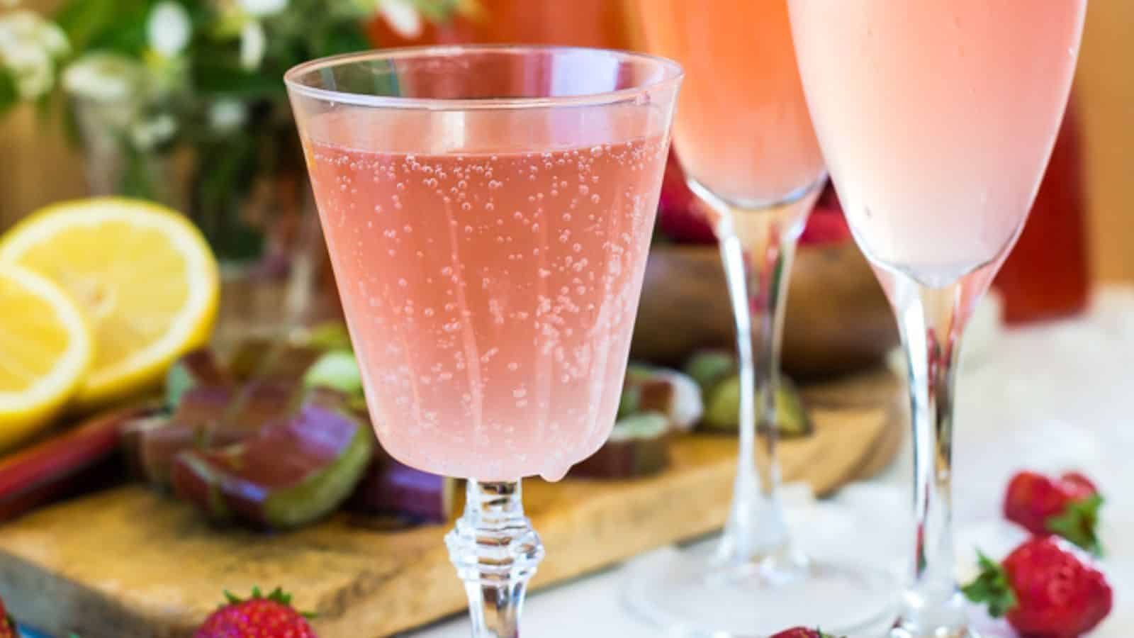 A glass filled with a bubbly pink drink is in focus. In the background, there are two more glasses with similar drinks. Cut lemons, strawberries, and flowers are also visible on a wooden board.