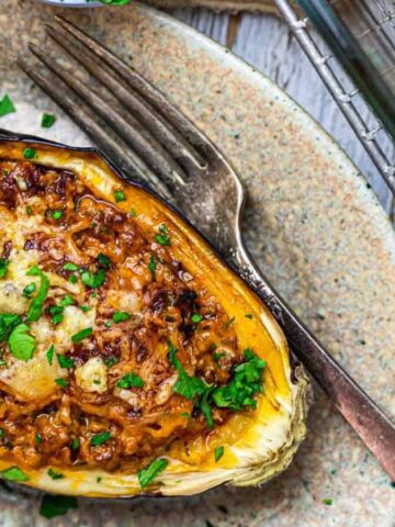 A stuffed roasted eggplant half garnished with chopped herbs on a speckled plate. A fork rests beside the eggplant. Another similar dish is partially visible on a wire rack.