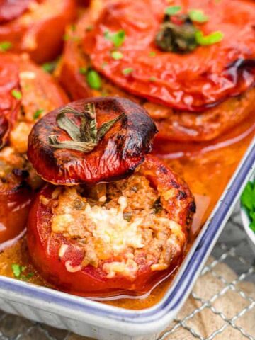 A baking dish filled with stuffed tomatoes, topped with herbs, resting on a wire rack. The tomatoes appear roasted, with slightly charred tops. A small bowl of fresh herbs is placed beside the dish.