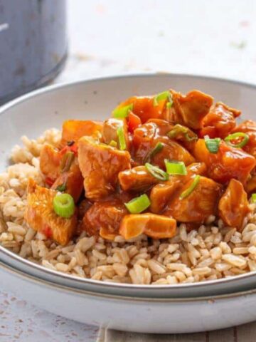 A bowl of brown rice topped with a saucy chicken and vegetable dish, garnished with chopped green onions. Behind the bowl is a slow cooker and part of a glass jar is visible. The scene is set on a light-colored surface with a striped cloth.