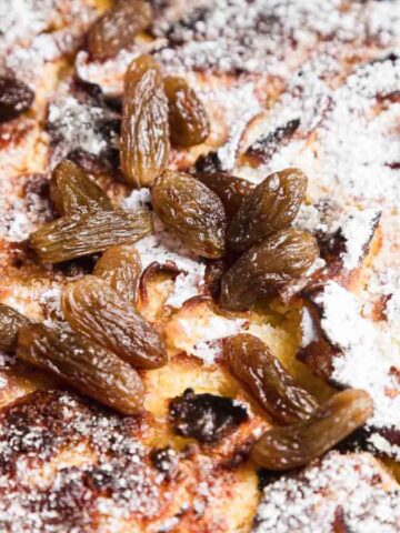Close-up image of golden-brown baked bread pudding topped with raisins and dusted with powdered sugar. The texture appears crispy and caramelized, with some darker edges visible. The raisins are scattered unevenly across the surface.