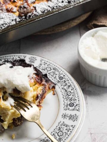 A slice of bread pudding on a decorative plate with a fork and a dollop of cream. In the background, part of a baking dish with additional bread pudding is visible. A small bowl of cream with a spoon rests beside it on a marble surface.