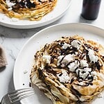 Two plates of roasted cabbage steaks topped with crumbled white cheese and drizzled with balsamic glaze rest on a marble surface. A fork lies beside them, while a dark bottle is partially visible in the background.
