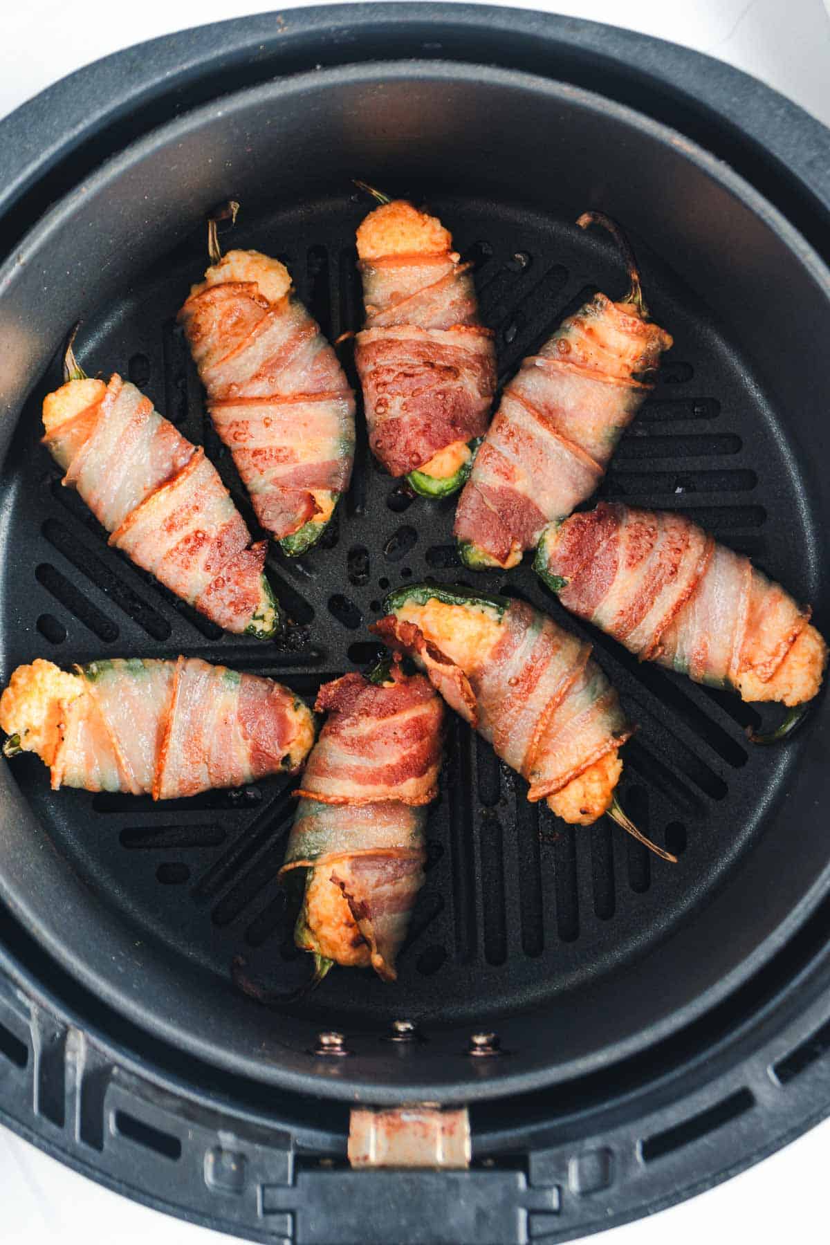 Eight air fryer jalape&ntilde;o poppers, wrapped in crispy bacon and stuffed with a savory filling, are arranged in a circular pattern inside the basket. The bacon is perfectly cooked with enticing grill marks enhancing each piece.