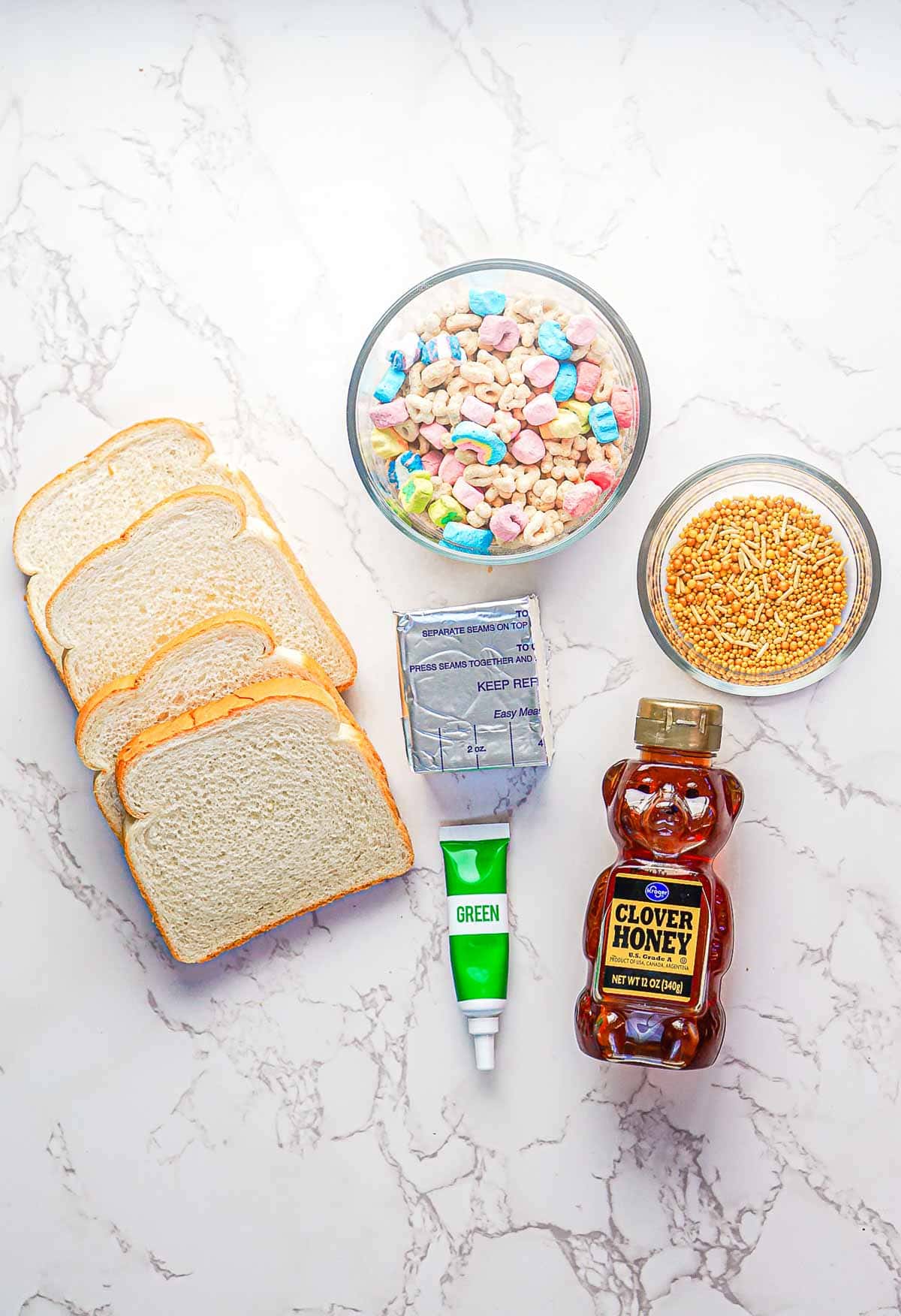 Slices of white bread, a tube of green icing, a package of butter, a jar of clover honey, a bowl of colorful marshmallow cereal, and a bowl of golden sprinkles are arranged on a marble surface.