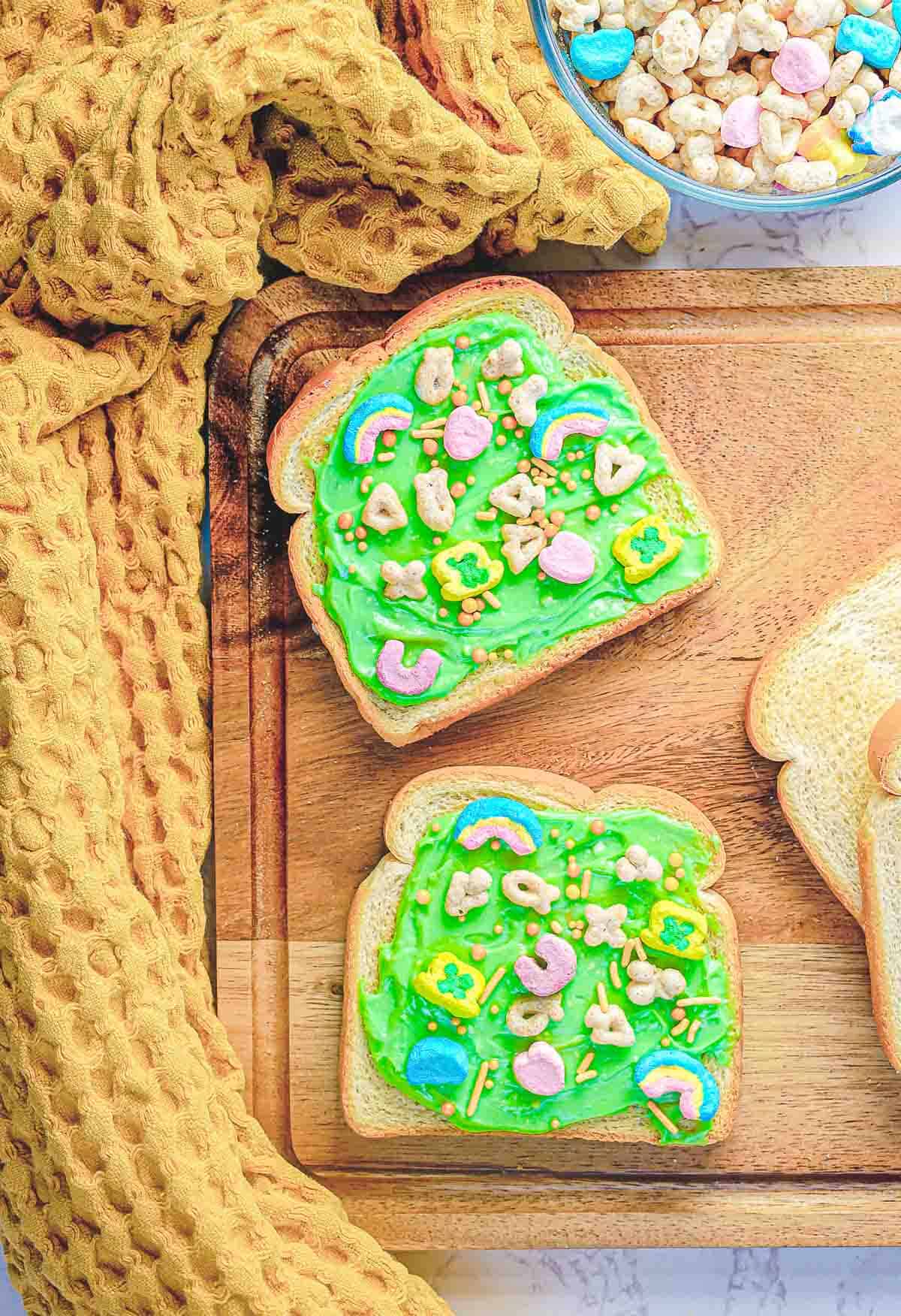 Two slices of Leprechaun Toast with green frosting and colorful cereal pieces rest on a wooden board. A textured yellow cloth is draped to the left, and a bowl containing similar cereal is partially visible at the top right, making it a festive breakfast perfect for St. Patrick's Day.