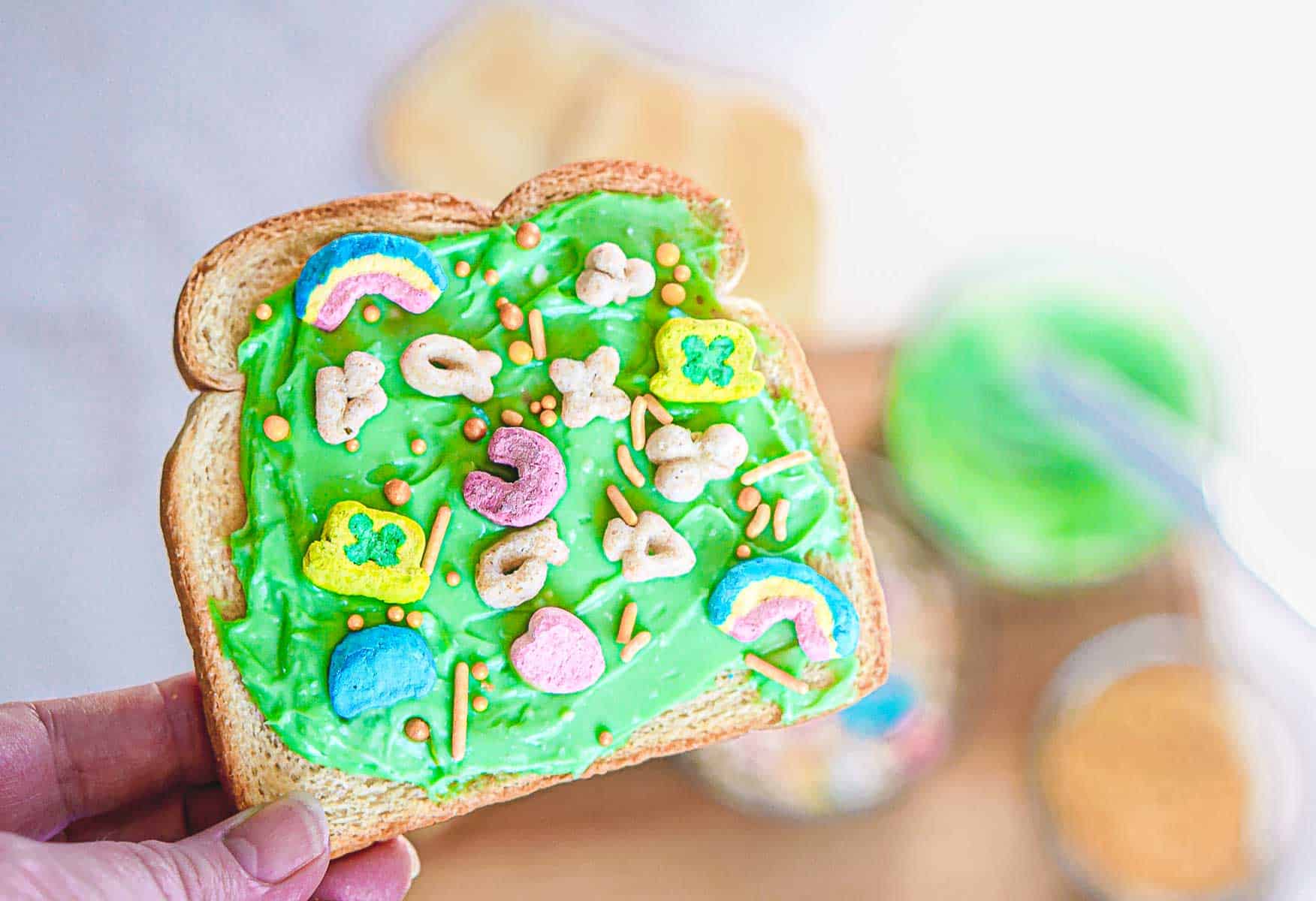 A slice of Leprechaun Toast with bright green frosting is topped with various colorful cereal pieces, including rainbows and clovers. The background shows a blurred mixing bowl with more of the St. Patrick's Day-inspired green frosting and a knife.