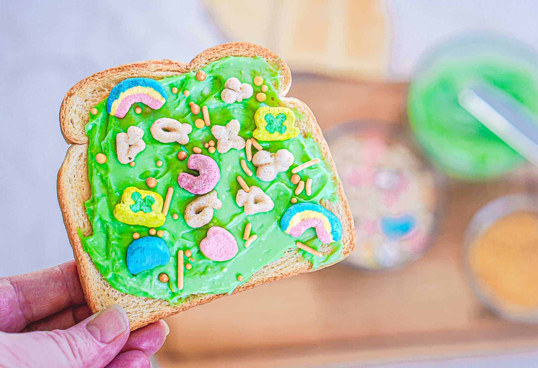 A hand holds a slice of Leprechaun Toast, spread with green frosting and topped with colorful cereal, like rainbows and stars. In the background, bowls brim with more cereal and toppings perfect for a whimsical St. Patrick's Day breakfast on a wooden surface.