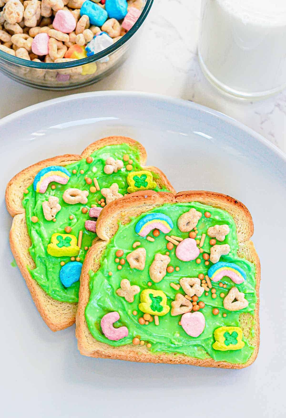 Start your morning with a festive breakfast treat: Leprechaun Toast. Two slices of toast with green frosting are topped with colorful cereal marshmallows and sprinkles, perfect for St. Patrick's Day. A bowl of cereal and a glass of milk add the finishing touch to this charming scene.