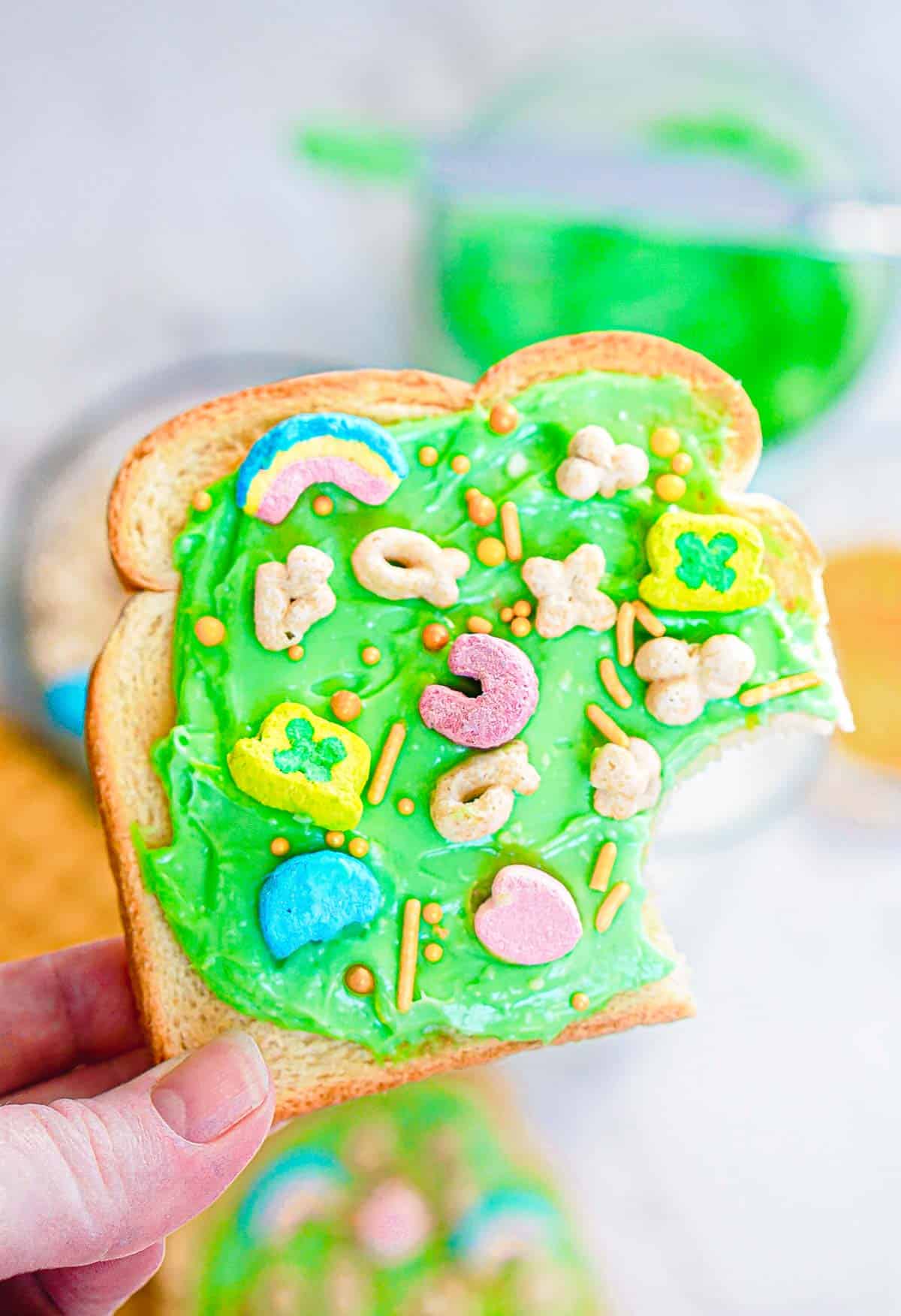 A slice of Leprechaun Toast spread with bright green frosting, topped with colorful marshmallow shapes and sprinkles. A bite is taken out of the bread. In the blurred background, a bowl of green frosting and another festive breakfast treat await their turn to celebrate St. Patrick's Day.