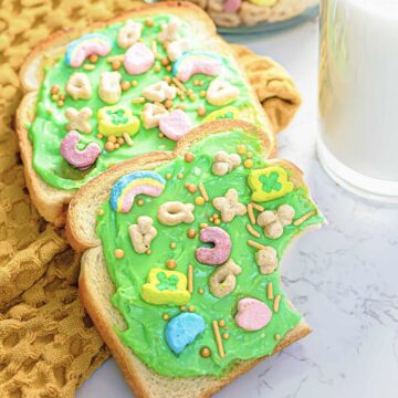 Two slices of Leprechaun Toast are spread with green frosting and topped with colorful cereal marshmallows and sprinkles. Festive and fun for St. Patrick's Day, one slice has a bite taken out of it. A glass of milk is visible nearby on a white marble surface, completing the whimsical breakfast scene.