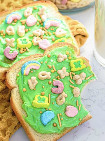 Two slices of Leprechaun Toast are spread with green frosting and topped with colorful cereal marshmallows and sprinkles. Festive and fun for St. Patrick's Day, one slice has a bite taken out of it. A glass of milk is visible nearby on a white marble surface, completing the whimsical breakfast scene.