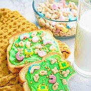 Two slices of bread spread with green frosting, topped with colorful cereal pieces and sprinkles, are placed on a yellow cloth. Behind them is a bowl of cereal and a glass of milk on a marble surface.