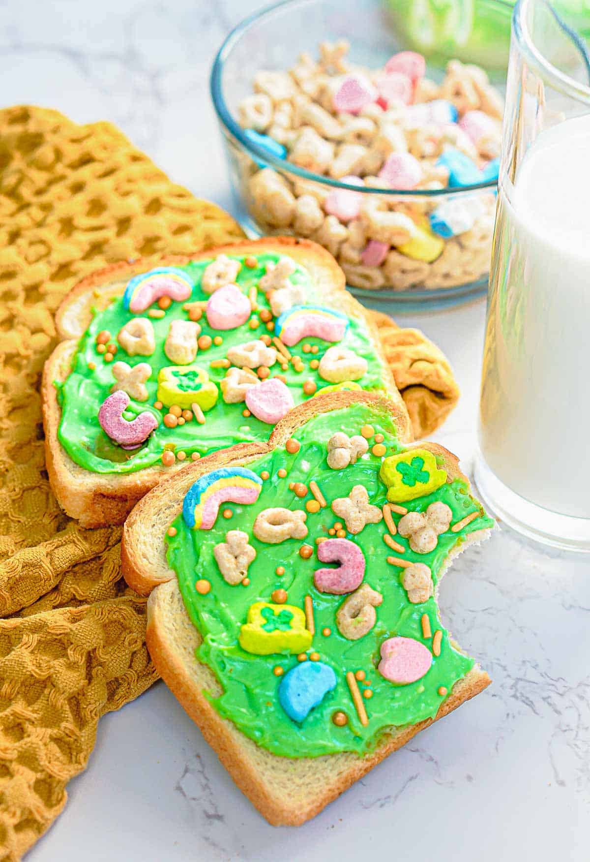 Two slices of bread spread with green frosting, topped with colorful cereal pieces and sprinkles, are placed on a yellow cloth. Behind them is a bowl of cereal and a glass of milk on a marble surface.