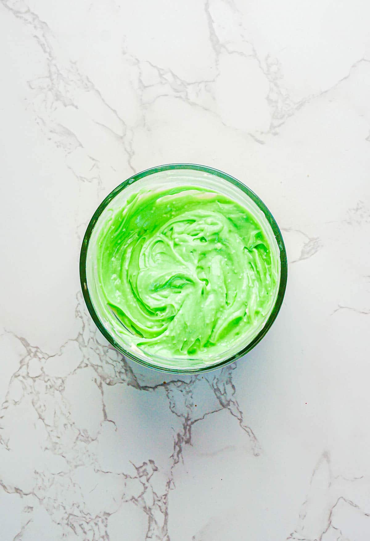 A glass bowl of creamy, light green frosting sits on a white marble counter, reminiscent of Leprechaun Toast ready for St. Patrick's Day festivities. The frosting appears smooth and whipped, with charming swirls on the surface.