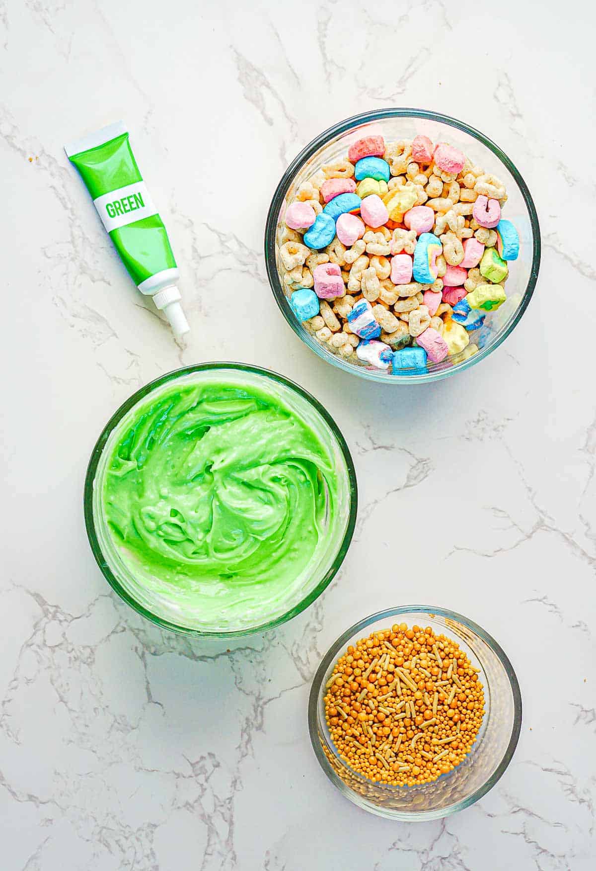 A tube of green food coloring lies next to a bowl of vibrant St. Patrick's Day frosting. Nearby, two small bowls brim with colorful marshmallows and cereal, perfect for Leprechaun Toast, while the other holds gold sprinkles, all beautifully arranged on a white marble surface.