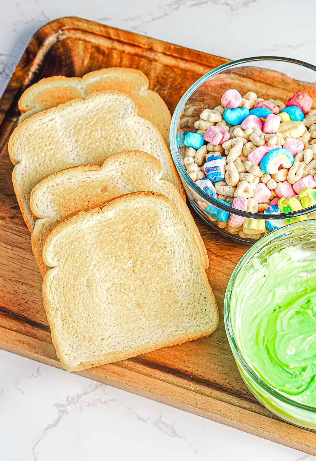 Slices of white bread, a bowl of cereal with colorful marshmallows, and a bowl of green-tinted spread are placed on a wooden tray for a whimsical St. Patrick's Day breakfast delight.
