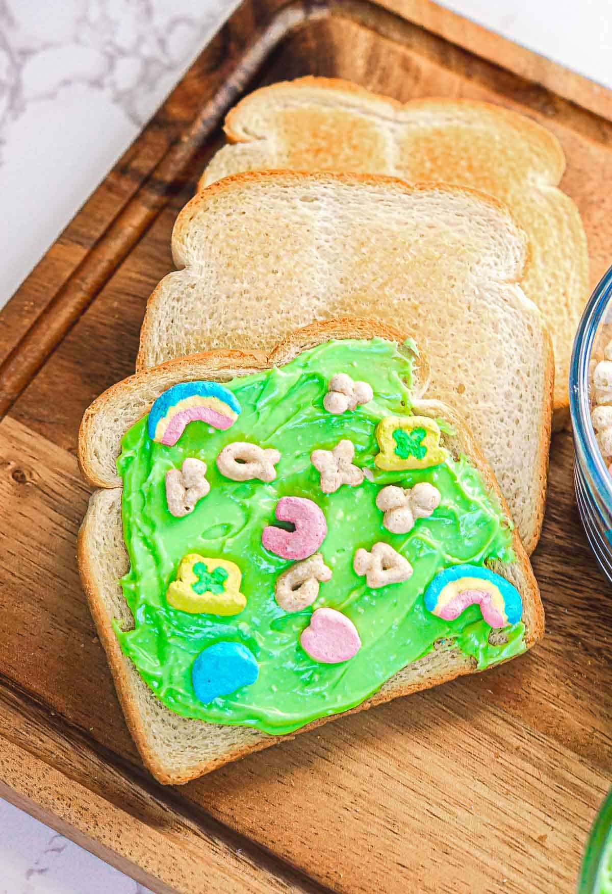 Slices of white bread on a wooden tray. The top slice, dubbed "Leprechaun Toast," is spread with green frosting and topped with colorful marshmallow pieces in rainbow, clover, heart, crescent, and star shapes—perfect for a festive St. Patrick's Day breakfast. A bowl of cereal sits on the right.